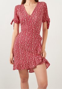 Robe florale rouge à manches courtes, col en V, boutons et ourlet volanté. Fabriquée en tissu léger avec une texture agréable et des accents de nœuds.