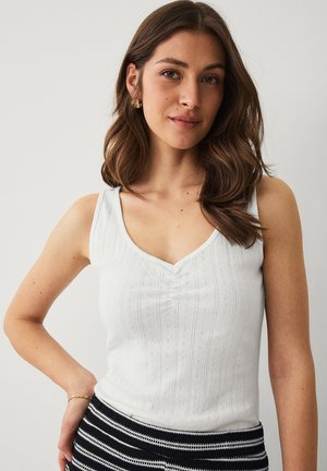 Jonge vrouw met golvend bruin haar, gekleed in een witte top zonder mouwen en een zwart-wit gestreepte rok, poseert met hand op de heup.