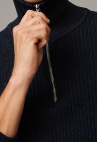 Pull tricoté bleu marine avec un col montant et une fermeture éclair en métal. La texture présente un motif côtelé, mettant en valeur un ajustement serré autour du cou.