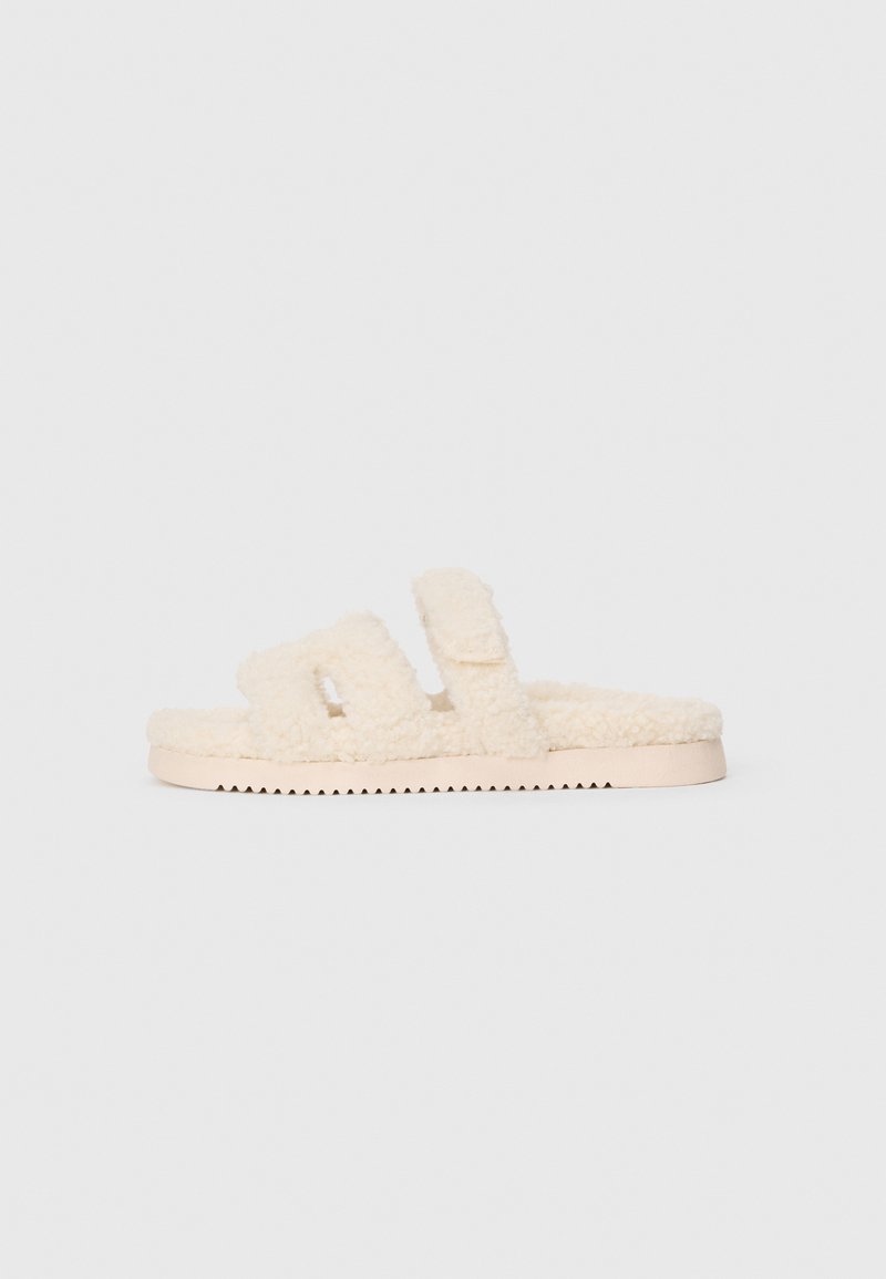 Cremefarbene, fuzzy Slide-Sandalen mit zwei breiten Riemen und flacher, geriffelter Sohle, seitlich auf weißem Hintergrund dargestellt.
