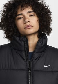 Μαύρο μπουφάν puffer με ψηλό γιακά, κλείσιμο με φερμουάρ και οριζόντια ραφή. Φέρει μικρό λευκό λογότυπο Nike στην περιοχή του στήθους.