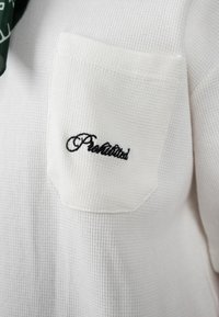 Tissu blanc texturé avec une petite poche poitrine brodée d'un texte noir en cursive indiquant « Prohibited ».