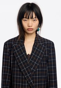 Granatowa marynarka w kratę z brązowymi i szarymi liniami siatki, szerokimi klapami i dopasowanym krojem. Noszona na ciemnym, koronkowym topie.