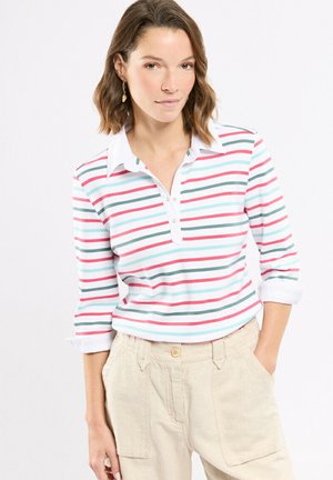Femme aux cheveux bruns mi-longs portant une chemise blanche à col avec des rayures horizontales rouges, vertes et bleues, et un pantalon beige, une main dans la poche.