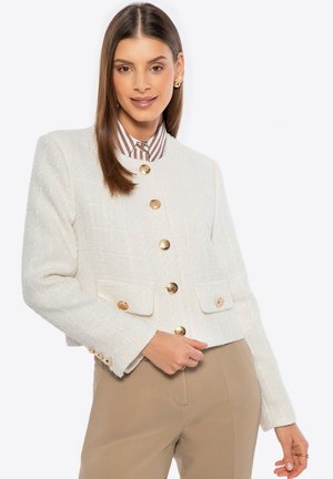 Witte tweed jasje met een gestructureerd ontwerp, gouden knopen en twee frontzakken. Afgewerkt met een afgeronde halslijn en textiel met structuur.