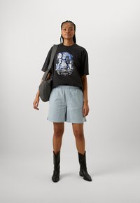 Black graphic t-shirt with blue design, light blue textured shorts, and black leather ankle boots. Model carries a gray checkered bag.