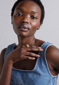 Denim top with wide shoulder straps, featuring a square neckline. Gold ring accent on hand; smooth skin texture and natural makeup.