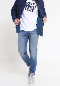 Giacca di jeans blu sopra una maglietta bianca con testo nero in grassetto. Indossata con jeans azzurri chiari e sneakers grigie. Sfondo semplice.