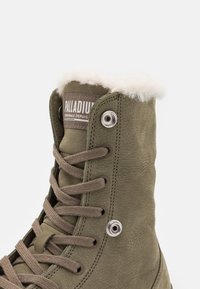 Botte montante vert olive avec lacets marron et bordure en fourrure blanche, avec œillets métalliques et un écusson logo "Palladium" sur la languette.