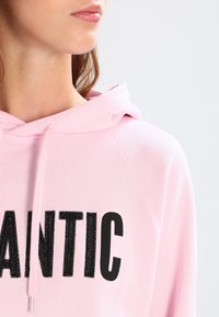 Sudadera rosa con letras texturizadas en negro que dicen "ANTIC." Presenta una capucha con cordón y un material suave similar al algodón.