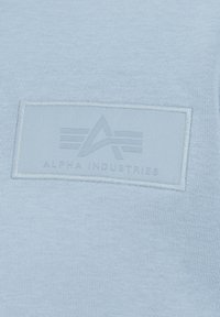 Svijetloplava tkanina dukserice s teksturiranim 'Alpha Industries' pečatom. Pečat prikazuje logotip s prugama i utisnutim dizajnom.