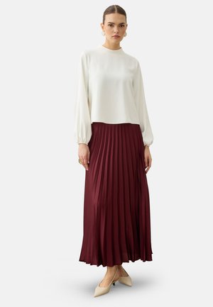 Donna che indossa una camicetta bianca a maniche lunghe, gonna maxi plissettata bordeaux, scarpe beige a punta con tacco basso e orecchini dorati.