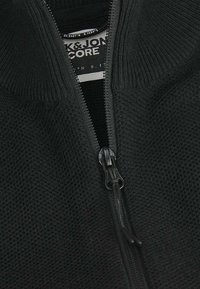 Veste zippée noire présentant un design en tricot texturé, un col côtelé et une doublure intérieure contrastante, avec une fermeture éclair en métal.