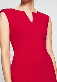 Robe rouge sans manches avec un décolleté en V et une coupe structurée. Présente des coutures en panneaux et une texture lisse, mettant en avant une silhouette cintrée.