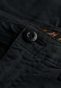 Nærbillede af sorte stofbukser med en sort knap med fire huller mærket "Superdry" og synlig orange stikning på linningen.