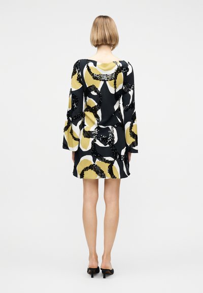 Γυναίκα με κοντά μαλλιά που φοράει μακρυμάνικο φόρεμα με ασπρόμαυρο και μουσταρδί αφηρημένο σχέδιο και μαύρα mules με ψηλά τακούνια, γυρισμένη από πίσω.