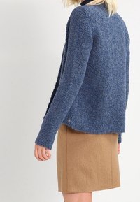 Suéter azul texturizado con mangas largas, frente abierto y un dobladillo trasero ligeramente más largo, combinado con una falda de lana beige.