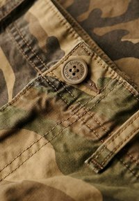 Tejido con estampado de camuflaje en tonos verdes y marrones, presentando un botón marrón con "SUPERDRY" grabado y detalles de costura visibles.