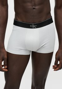 Άνδρας που φοράει λευκά μποξεράκια Calvin Klein με μαύρη μέση που δείχνει το λογότυπο της μάρκας, στέκεται με τα χέρια χαλαρά στα πλάγια.