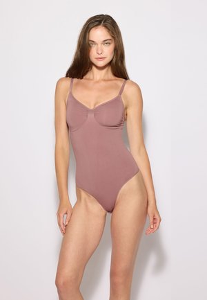 Giovane donna con lunghi capelli castani che indossa un body intero color malva, in piedi davanti a uno sfondo chiaro e uniforme.