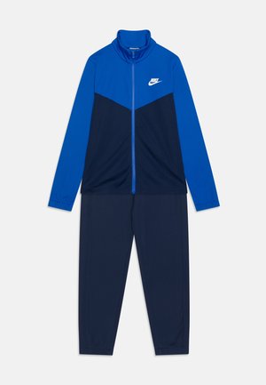 Modrý a námořnický teplákový komplet s zipovou bundou, vysokým límcem a dlouhými rukávy. Na hrudi je logo Nike. Materiál se zdá být hladký a sportovní.