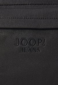 Črna tkanina z teksturirano površino, ki vsebuje reliefni logotip "JOOP! JEANS". Oblikovanje vključuje subtilne detajle šivanja.