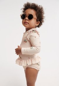 Maillot de bain rose pâle à motif floral, avec manches longues, détails à volants et bas assorti. Lunettes de soleil rondes teintées ajoutent une touche de style.