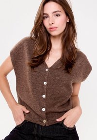 Cardigan en tricot marron avec un col en V, des manches courtes et cinq boutons blancs sur le devant. Texture lisse, coupe décontractée et style casual.