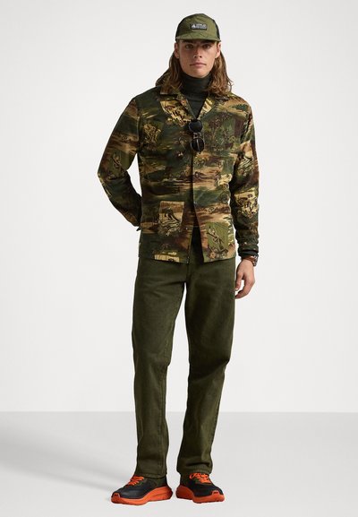 Cămașă cu model camuflat în nuanțe de verde și maro, cu nasturi; pantaloni oliv; adidași negri cu accente portocalii. Căciulă cu logo, ochelari de soare purtați.
