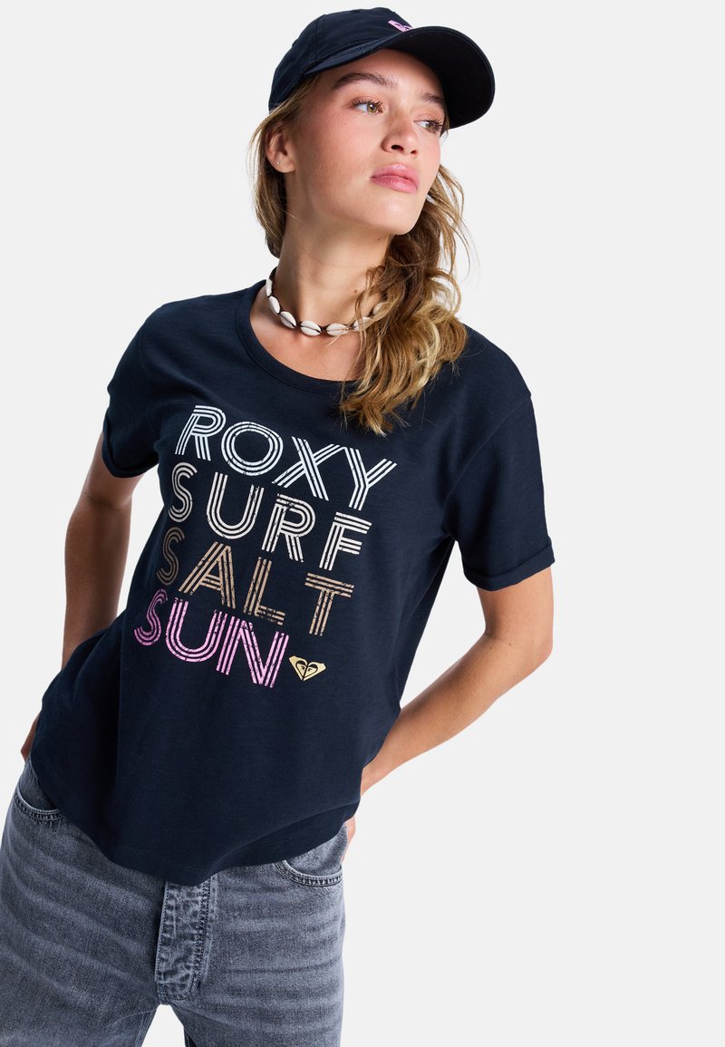 Young woman wearing a navy cap, shell necklace, navy T-shirt with "ROXY SURF SALT SUN" text, and faded jeans, posing with hands in pockets.