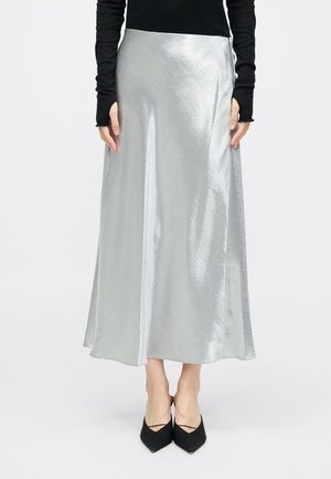 Jupe maxi en satin argenté avec une texture lisse, présentant une fente sur le côté. Portée avec un haut ajusté noir à manches longues et des ballerines noires à bout pointu.