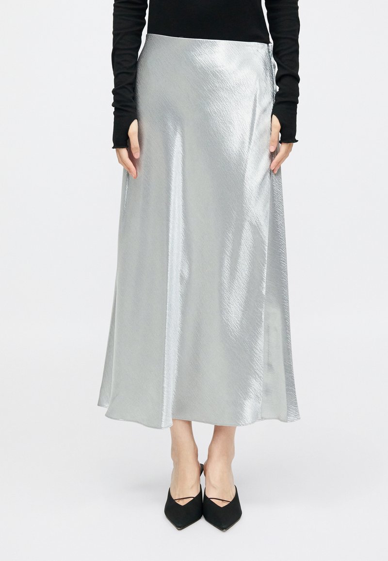 Srebrna satenska maxi krila z gladko teksturo, ki ima stranski razporek. Uporabljena je z oprijetim črnim dolgim rokavom in črnimi pointed balerinkami.