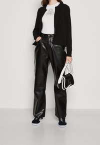 Czarna długa kurtka z długim rękawem na białą koszulę z grafiką, zestawiona z czarnymi skórzanymi spodniami o szerokich nogawkach i czarnymi sneakersami. Zestaw uzupełnia mała czarno-biała torebka.