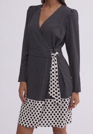 Kobieta ubrana w czarną i białą sukienkę kopertową w groszki z długimi rękawami i kontrastującym paskiem w talii.