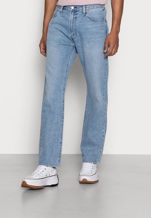 Homme portant un jean droit bleu clair et des baskets blanches à plateforme, debout sur un sol gris devant un mur blanc.