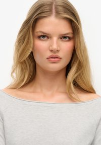 Hellgraues, schulterfreies Oberteil mit weichem, glattem Material und Raglanärmel. Das Model hat glattes, langes, blondes Haar und dezentes Make-up.