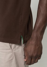 T-shirt en coton marron avec une fente latérale et une couture accentuée verte, associé à un pantalon beige clair, mettant en valeur le tissu texturé.