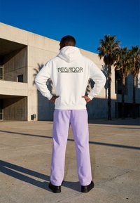 Homme debout en plein air, les mains sur les hanches, portant un sweat à capuche blanc avec le texte « PEGADOR Moments & memories » et un pantalon violet clair sous un ciel bleu clair.