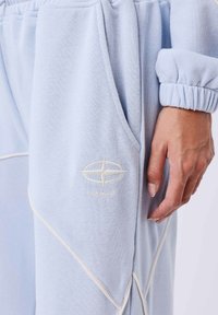 Lichtblauwe joggingbroek met witte stikseldetails, zijzak, elastische tailleband en boorden, en een hand die naast de zak rust.