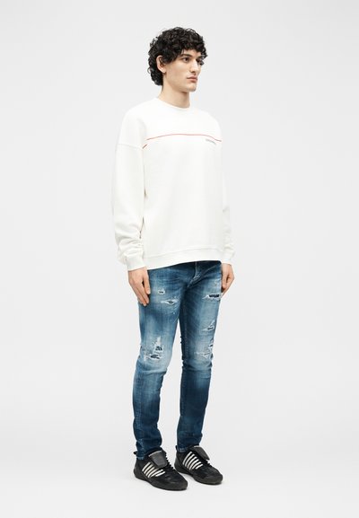 Νέος άντρας με σγουρά μαλλιά φορώντας λευκό φούτερ, σκισμένα μπλε τζιν και μαύρα αθλητικά, που στέκεται μπροστά σε απλό φόντο.