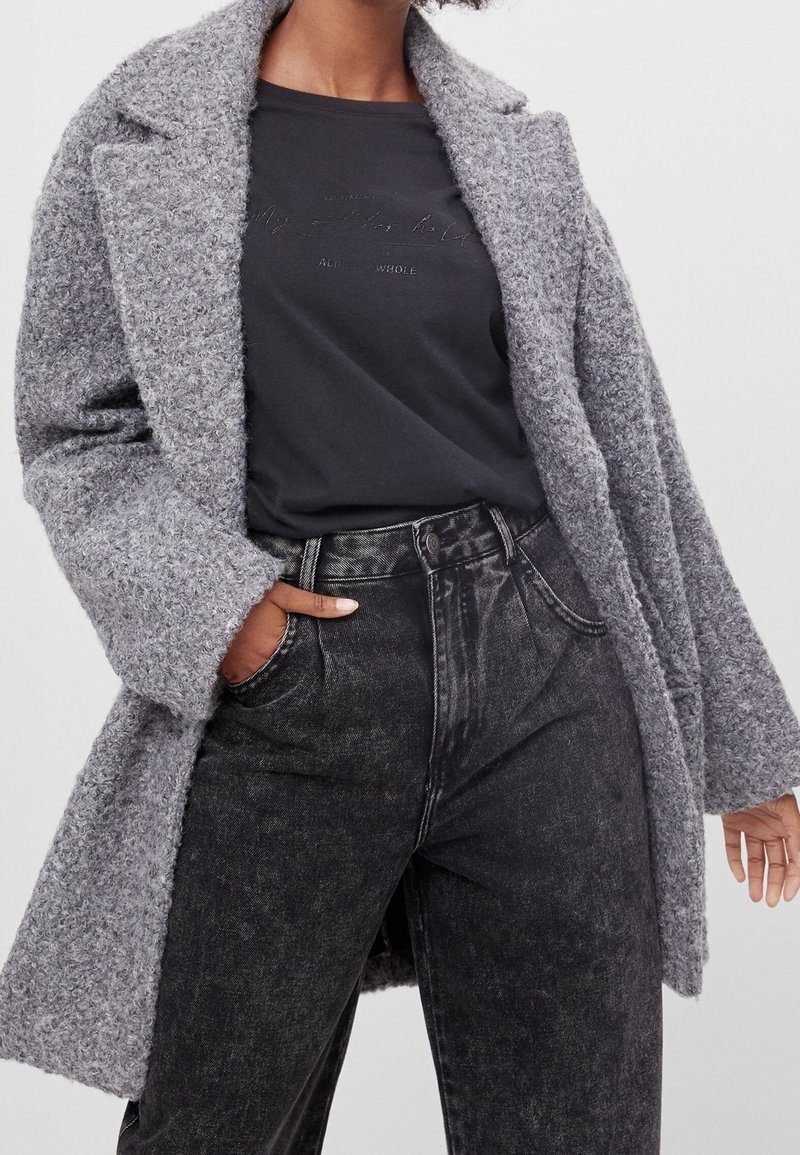 Abrigo largo gris texturizado con solapas muesca sobre una camisa negra y unos jeans de tiro alto gris y negro. Los detalles notables incluyen mangas anchas y bolsillos.