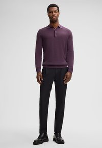 Uomo in piedi che indossa un polo tricot a maniche lunghe viola, pantaloni neri su misura e scarpe eleganti in pelle nera, su uno sfondo grigio uniforme.