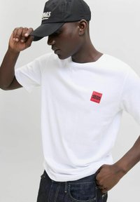 Hvid bomulds t-shirt med rund hals og korte ærmer, med et lille rødt firkantet logo på venstre bryst. Mørke jeans og en sort cap.