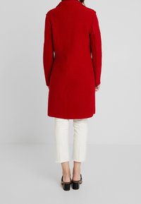 Röd ullkappa med rak design, långa ärmar, krage och längd till mitten av låren. Har sidofickor och bärs över vita, korta byxor.