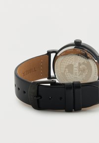 Montre avec bracelet en cuir noir, boîtier rond, dos transparent affichant un motif de globe, et boucle en métal. Résistante à l'eau jusqu'à 50 mètres.