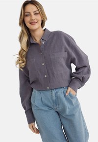 Fioletowa koszula z guzikami z przodu, posiadająca dwie kieszenie, wykonana z teksturowanej tkaniny, w połączeniu z jasnoniebieskimi, luźnymi dżinsowymi spodniami.