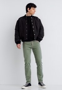 Νέος άνδρας που φοράει μαύρο μπουφάν μπόμπερ, μαύρο πουκάμισο, πράσινο παντελόνι και μαύρα αθλητικά παπούτσια, στέκεται μπροστά σε ένα λευκό τοίχο κοιτάζοντας προς τα αριστερά του.