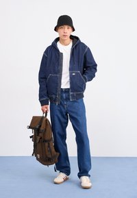 Denimjakke med hætte, blå med kontraststing, parret med blå jeans. Mørkebrun rygsæk med justerbare stropper holdt i den ene hånd.