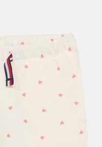 Παντελόνια με ελαστική μέση σε εκρού ύφασμα με μικρά ρόδινα σχέδια καρδιών· διαθέτουν ριγωτή λεπτομέρεια σε κόκκινο, λευκό και μπλε.
