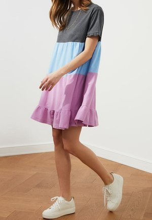 Vrouw loopt en draagt een korte jurk met kleurvlakken in grijs, blauw en lavendel, gecombineerd met witte sneakers op een houten vloer.